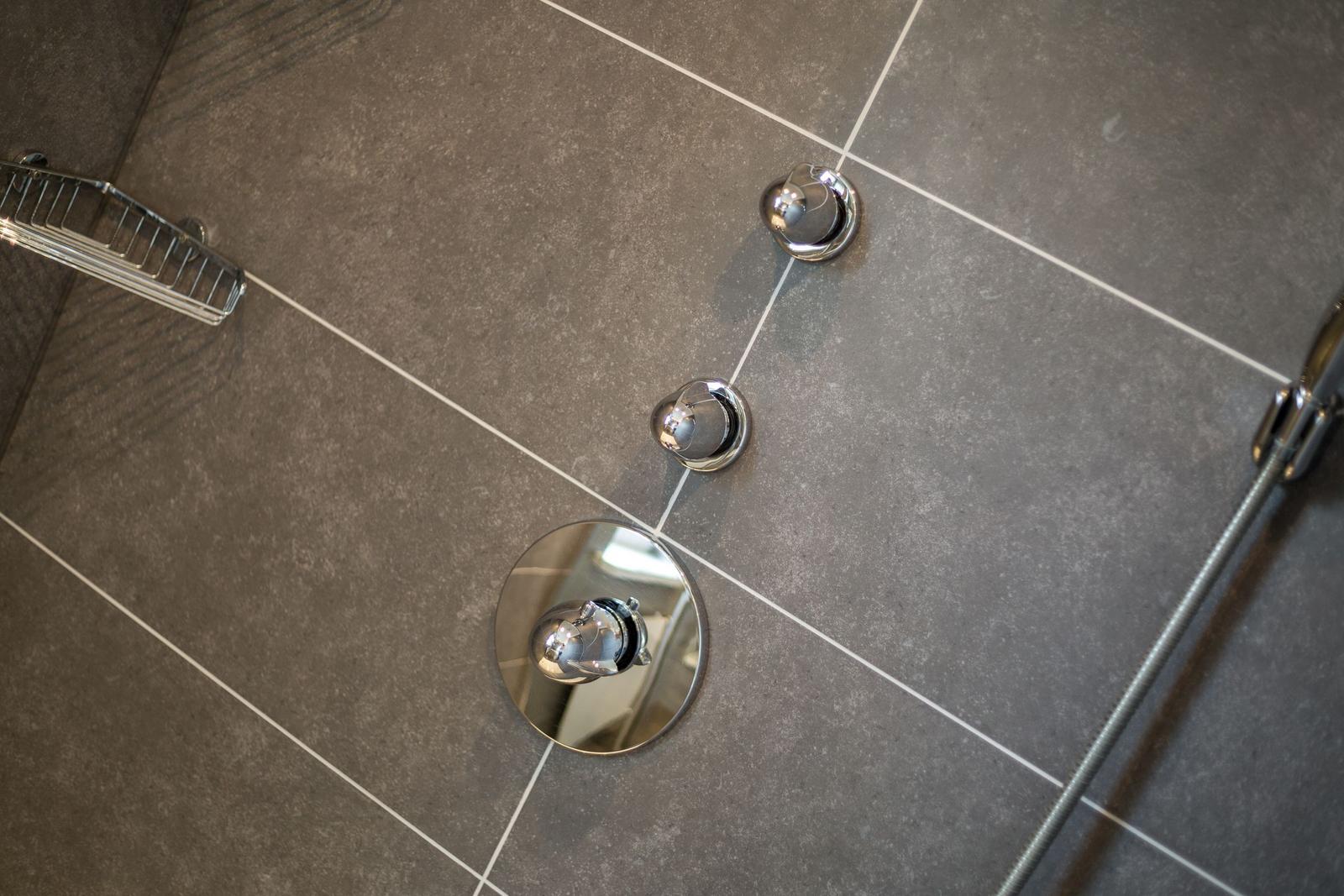 Duschkopf und Bedienelemente auf grauem Fliesenboden mit silbernen Griffen.