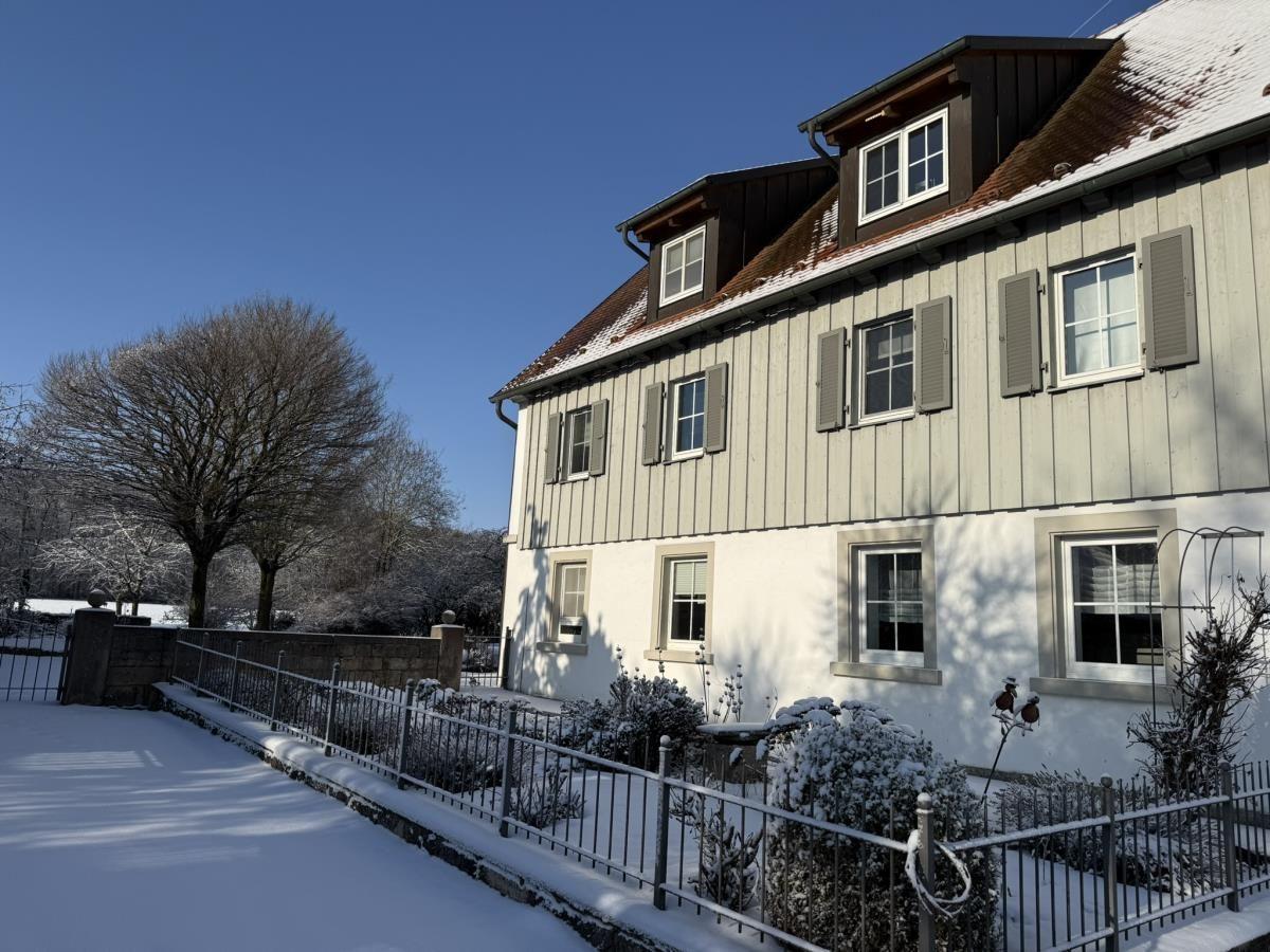 Wohnhaus mit Schneebedecktem Dach und Fenstern, umgeben von schneebedeckten Sträuchern und einem Zaun.