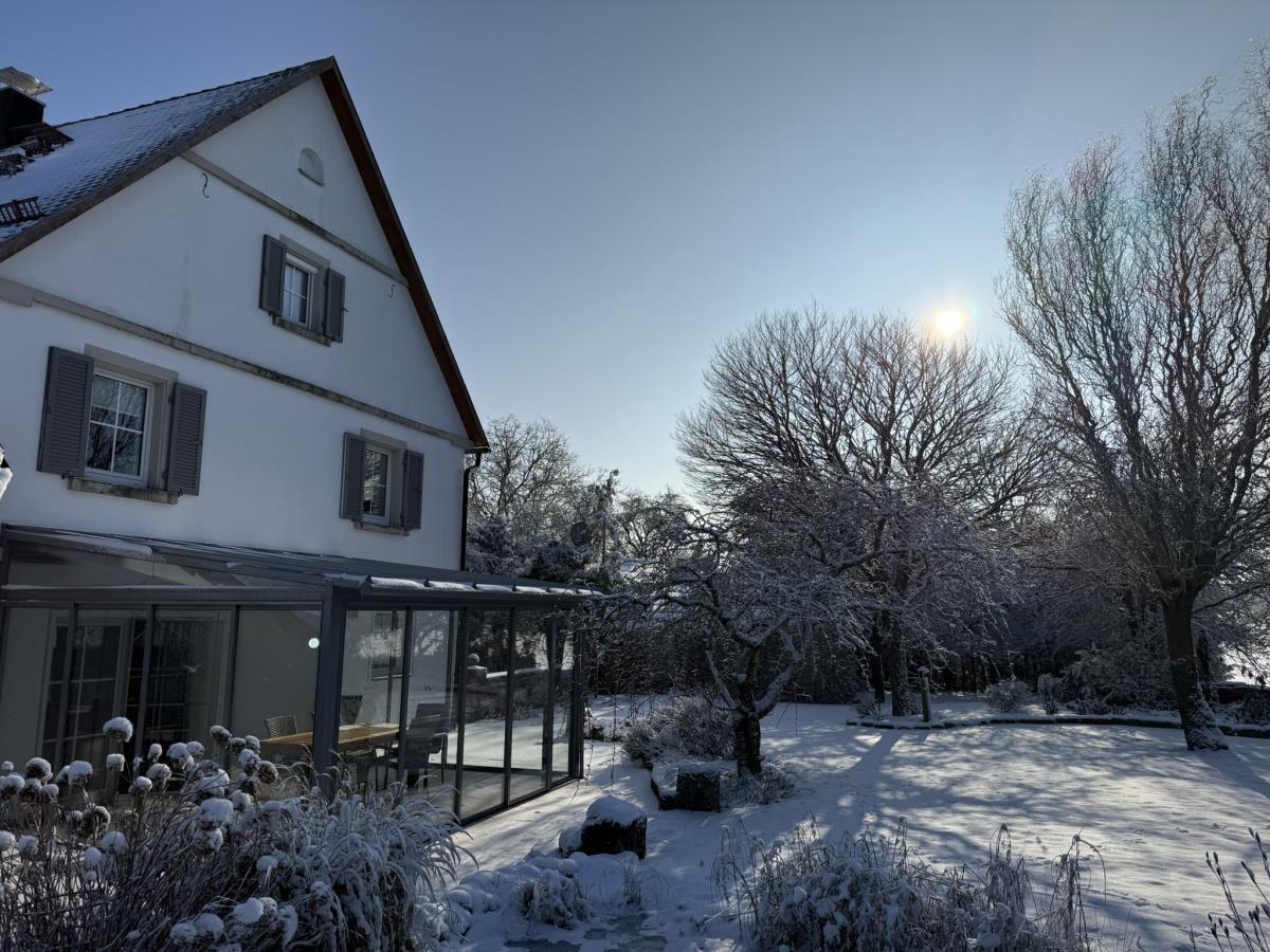Schneebedecktes Haus mit Wintergarten und schneebedeckten Bäumen im Garten.