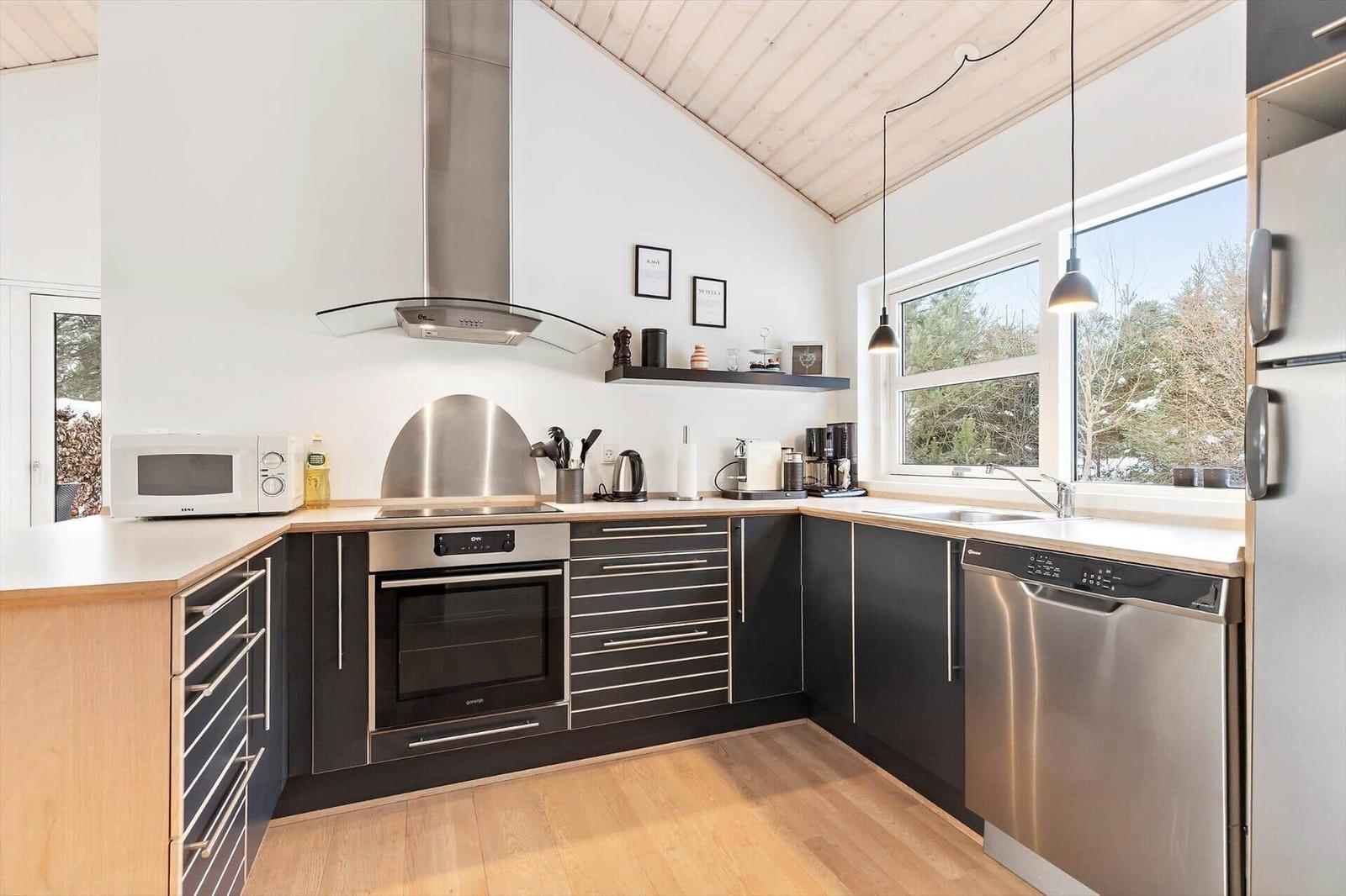 Kitchen with black cabinets, stainless steel appliances, and wooden floor. Windows with view of forest.