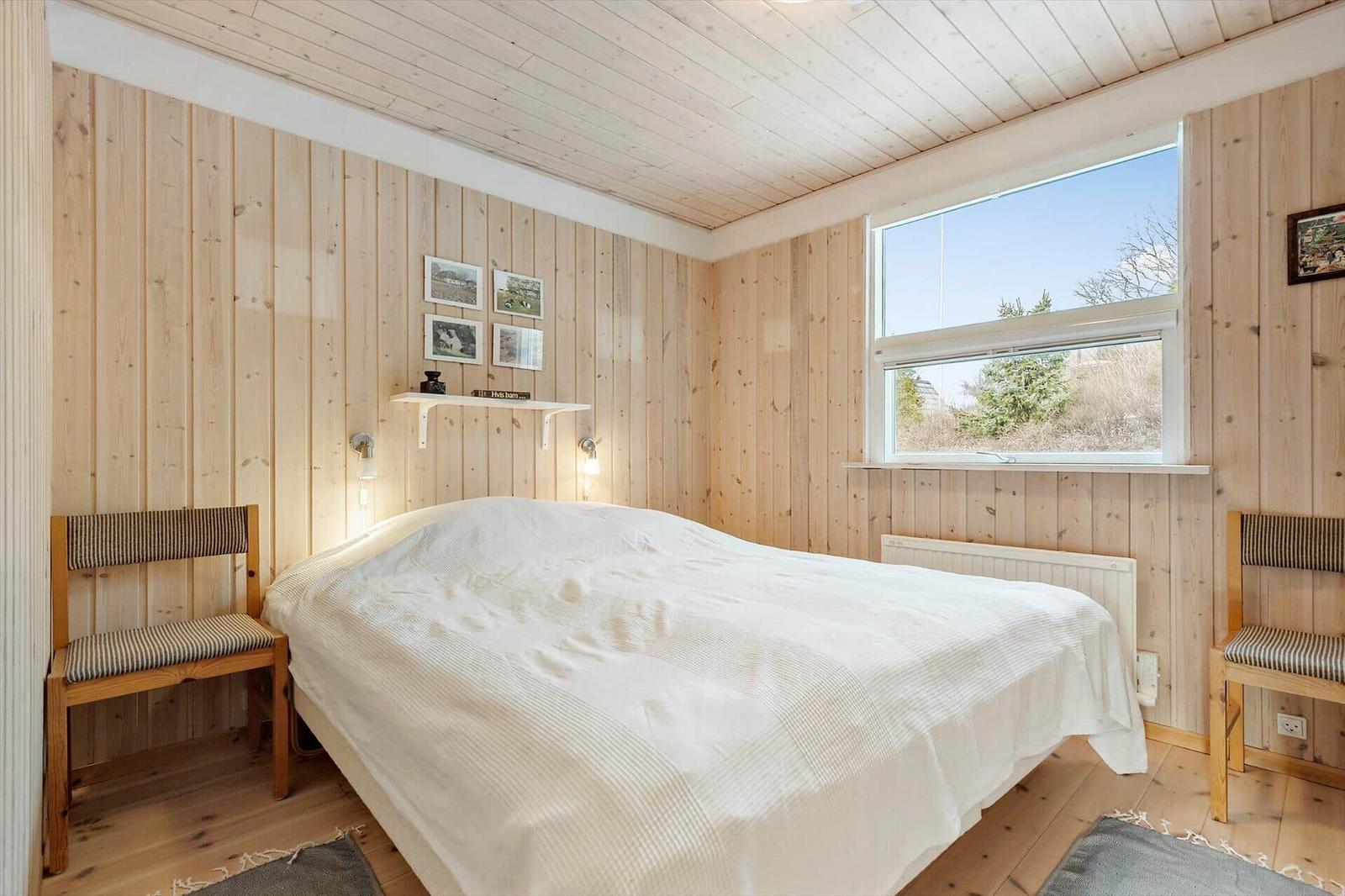 A bedroom with wooden walls, bed, two chairs, and window. 