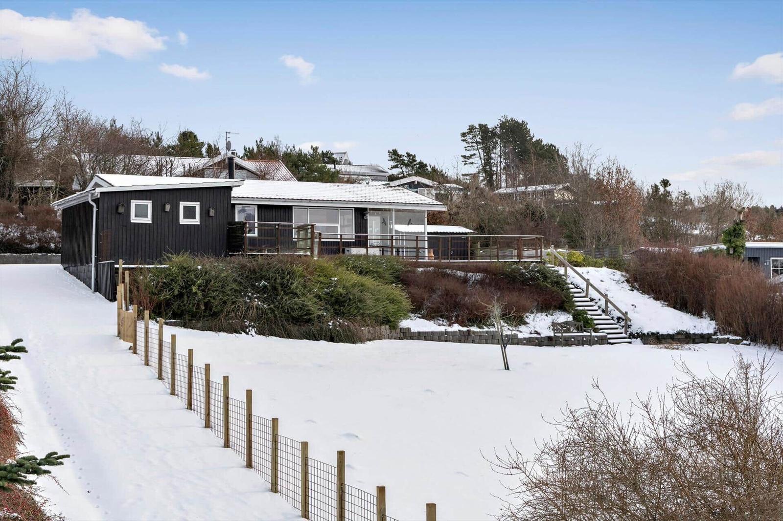 Black house with terrace and snow-covered garden. Wooden railings and steps lead to the entrance.