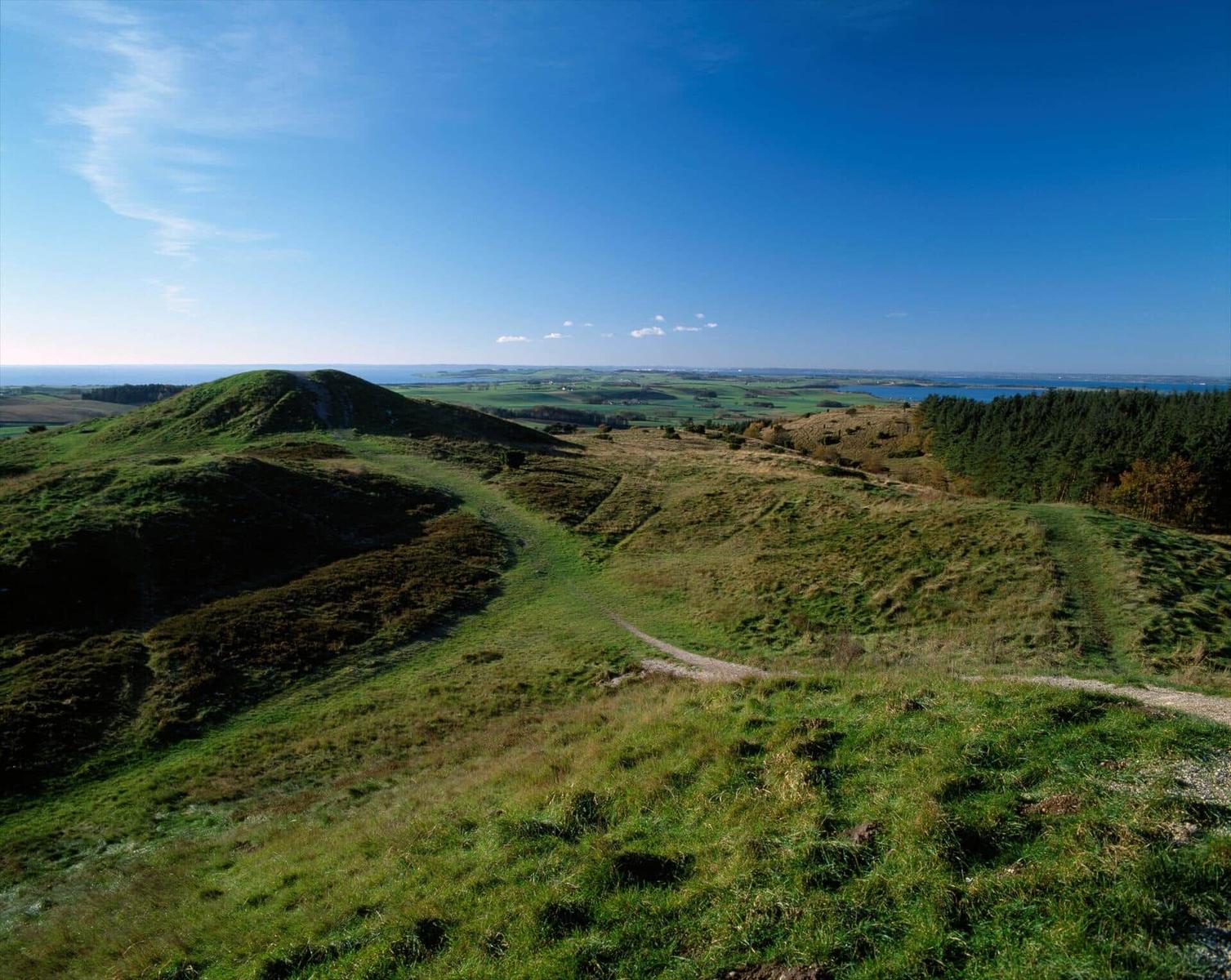 Green hills with a hiking trail and view of the sea under a blue sky.