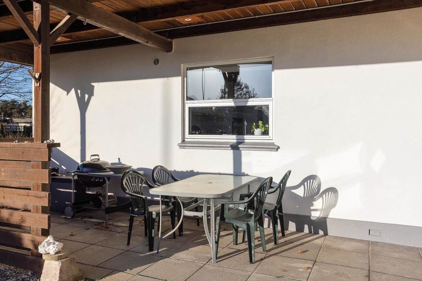 Terrace with table, chairs, and grill under wooden cover. Window with plant on sill.