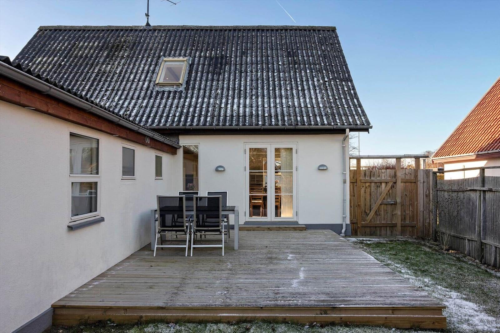 House with wooden terrace, garden, and garden gate. Outdoor table with chairs. Roof with skylight.