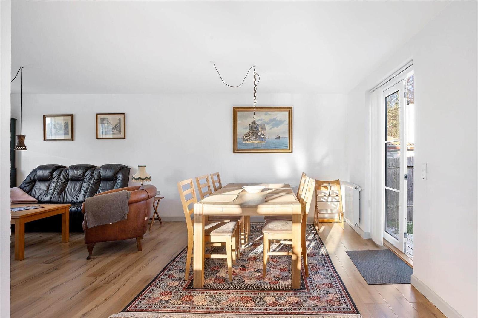 Dining room with wooden table, chairs, and rug. Living area with sofa and armchair. Window to outdoor terrace.