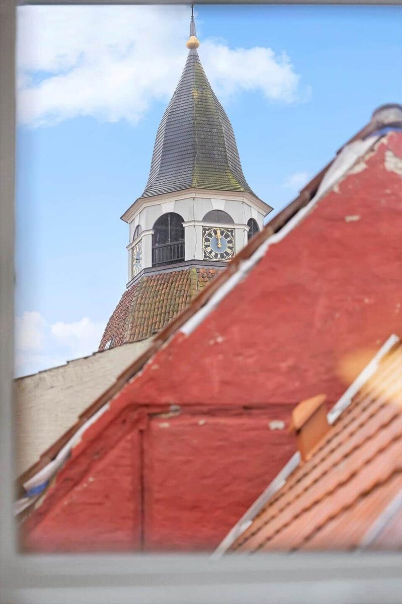 From the window, a church tower with clock and pointed roof is visible, surrounded by red roofs.