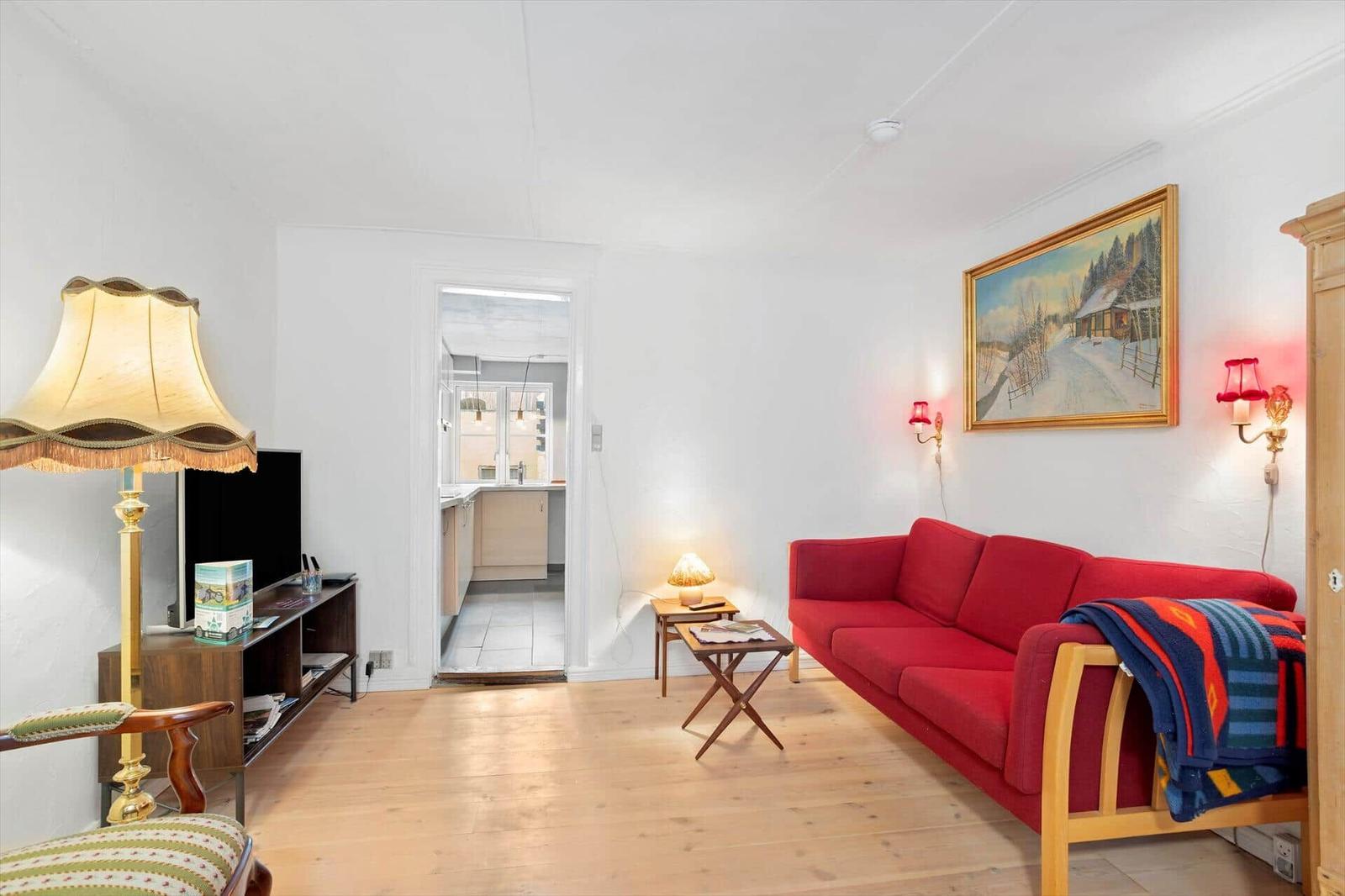 A living room with a red sofa, wooden floor, and view into the kitchen.
