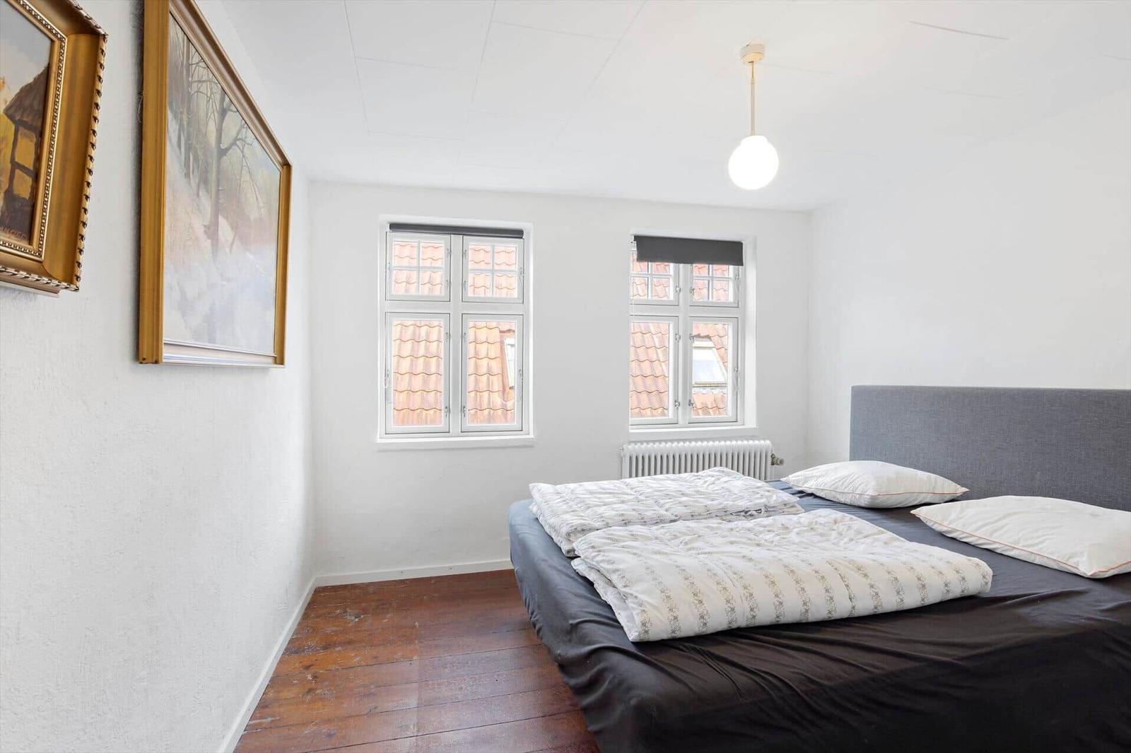 Bedroom with bed, two windows, and wooden floor. Two pictures hang on the wall.