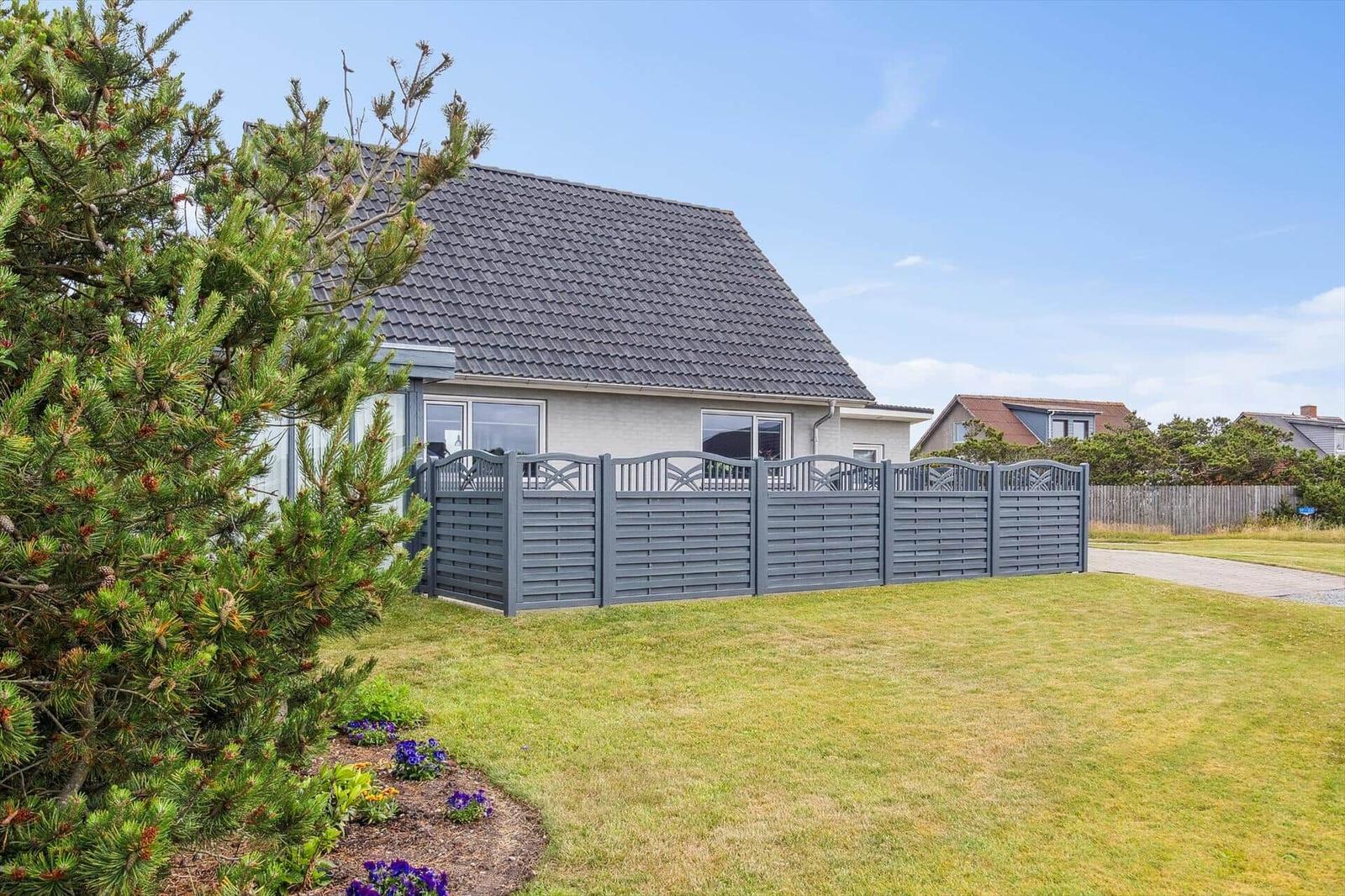 Haus mit grauem Dach und Zaun auf grünem Grundstück unter blauem Himmel.