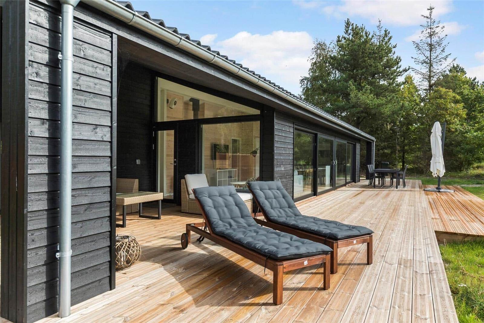 Deck with lounge chairs, table and chairs. Wood siding and glass facade. Background: forest.
