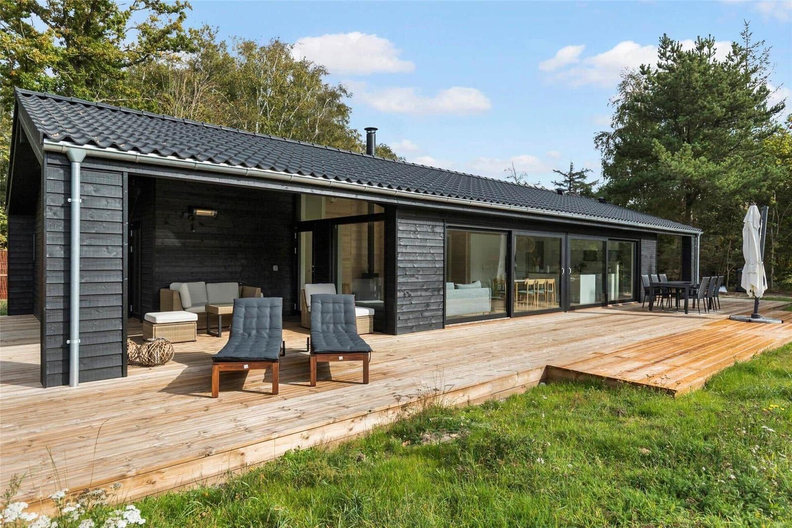 Modern wooden cabin with large wooden terrace, lounge area, and view into the forest.