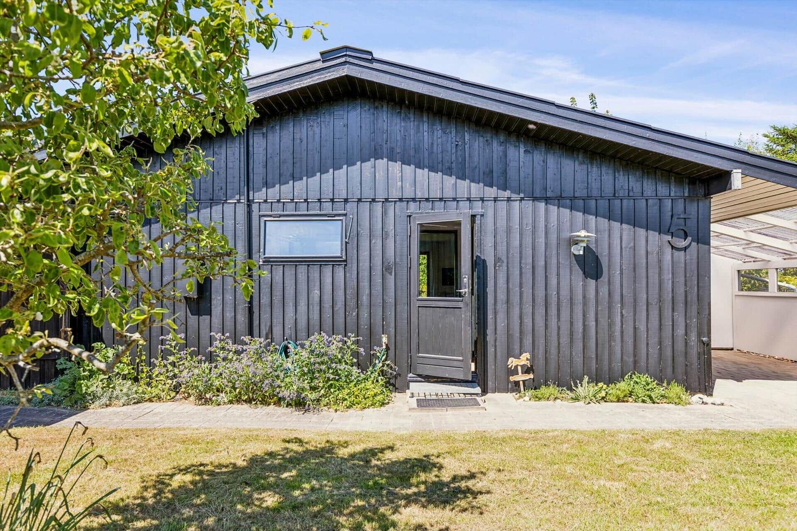 Black wooden house with entrance door and small window. Garden area with flowers and trees.