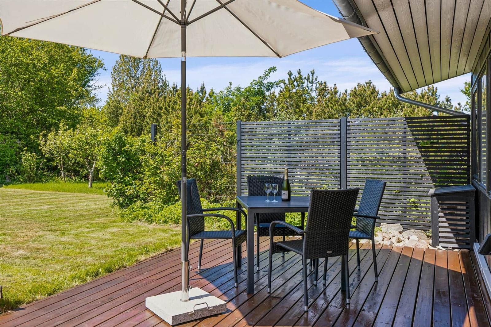 Deck with table, four chairs, and umbrella. Background: green lawn and wooden railing.