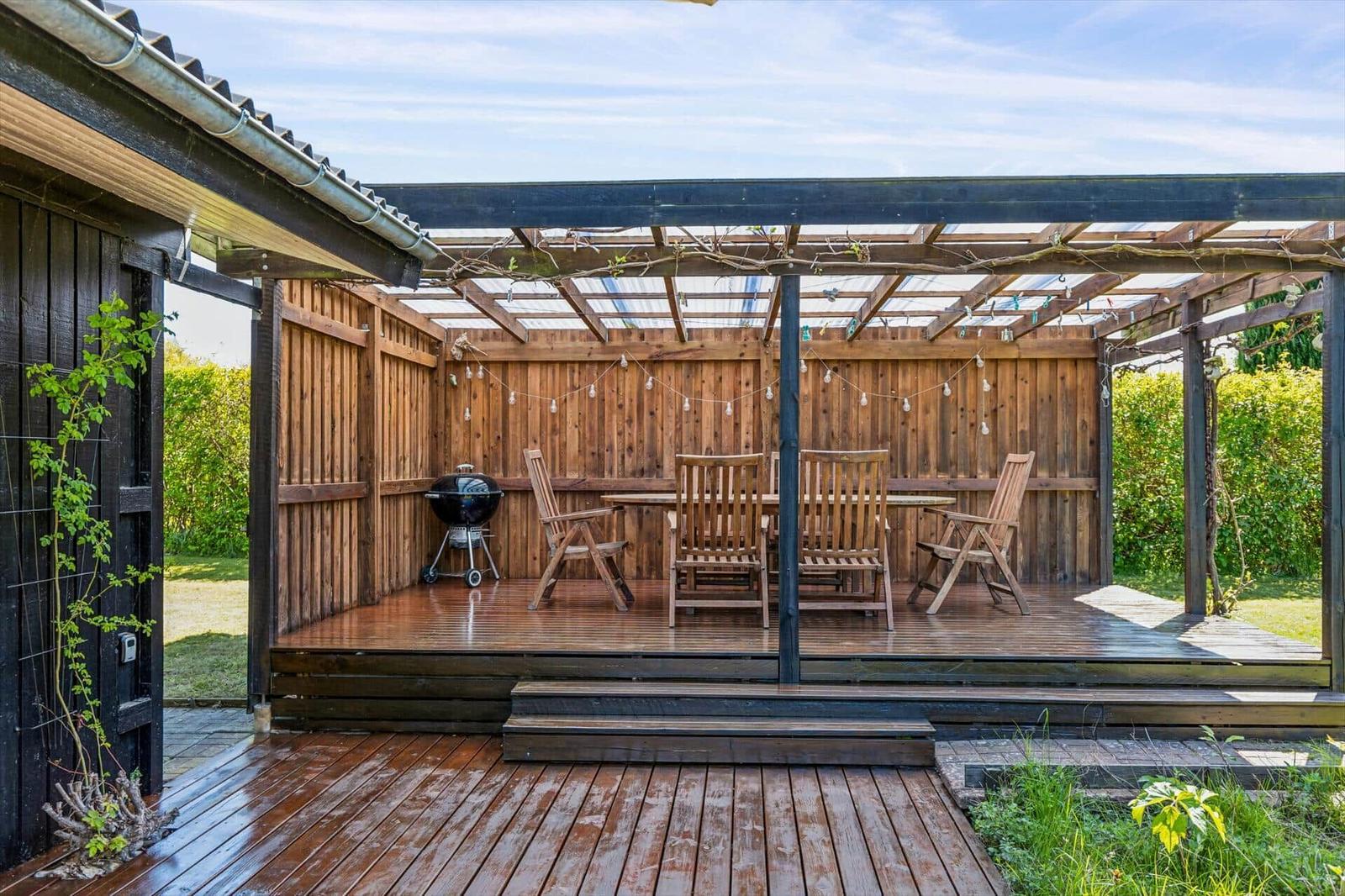 Wooden terrace with seating, grill, and pergola under a roof.
