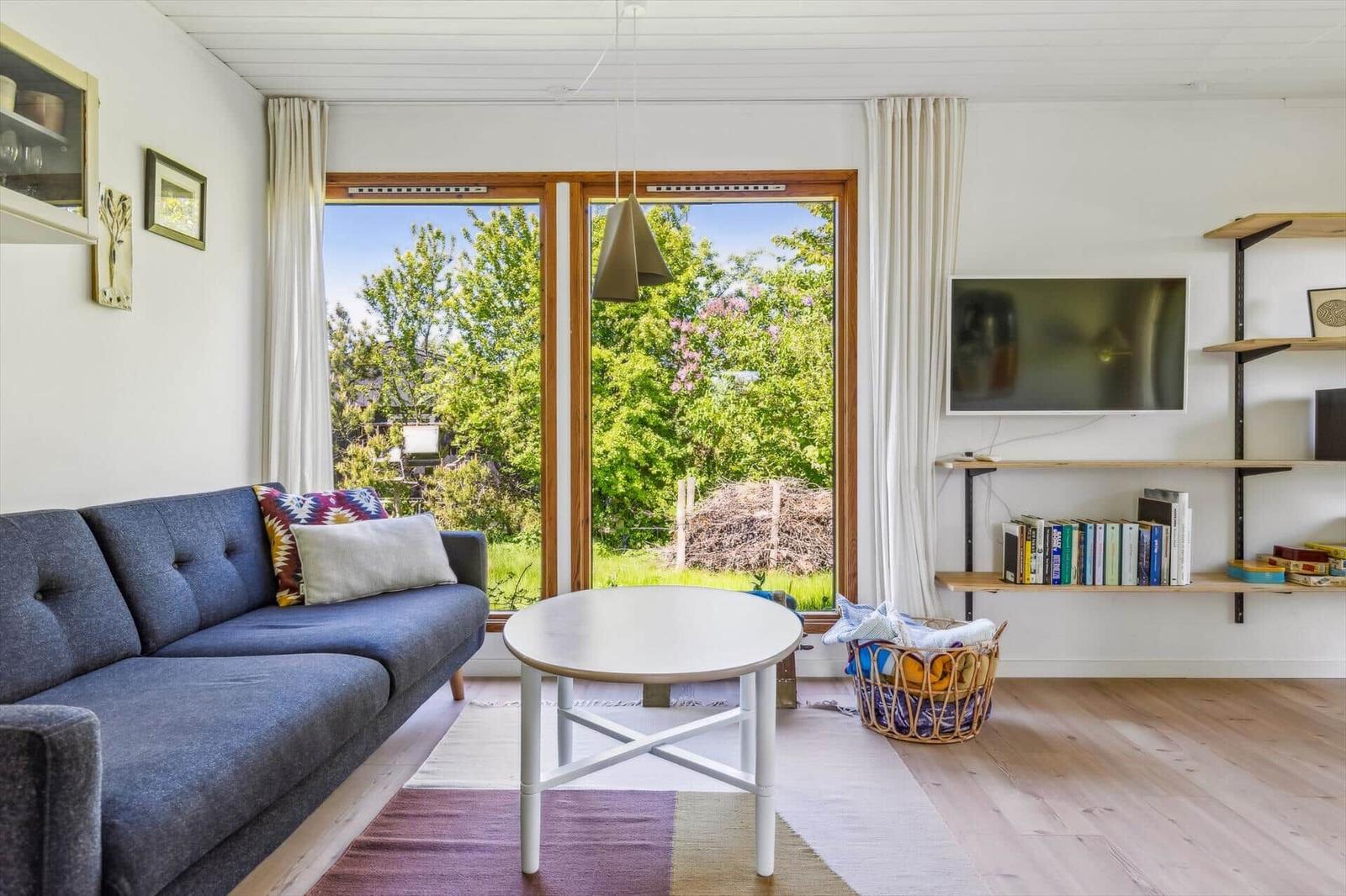 Living room with sofa, table, and TV. Green view through windows.