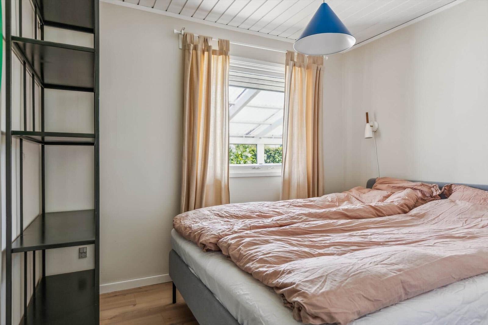 Bedroom with bed, window, and wall lamp. Visible wooden floor and metal shelf.