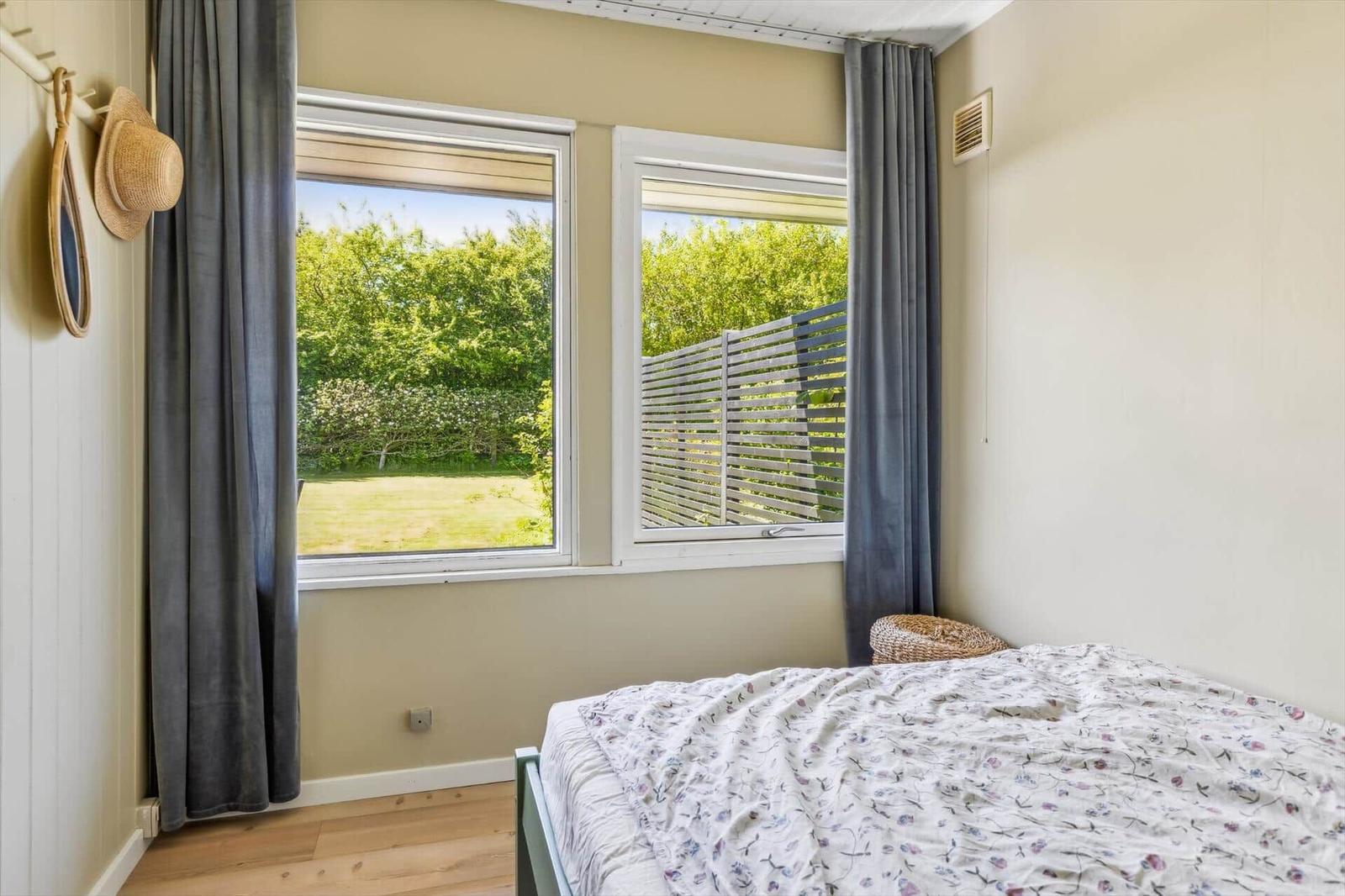 Bedroom with view of green garden and wooden terrace. Bed, curtains, and wooden floor.