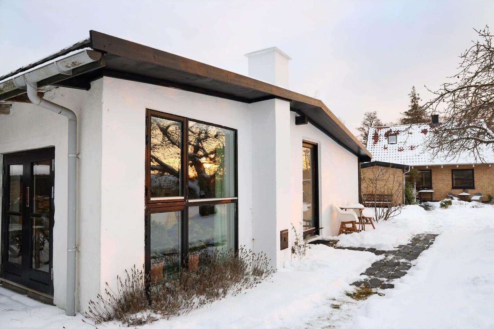 A white house with large windows and a snow-covered garden. A terrace with a table and chairs.