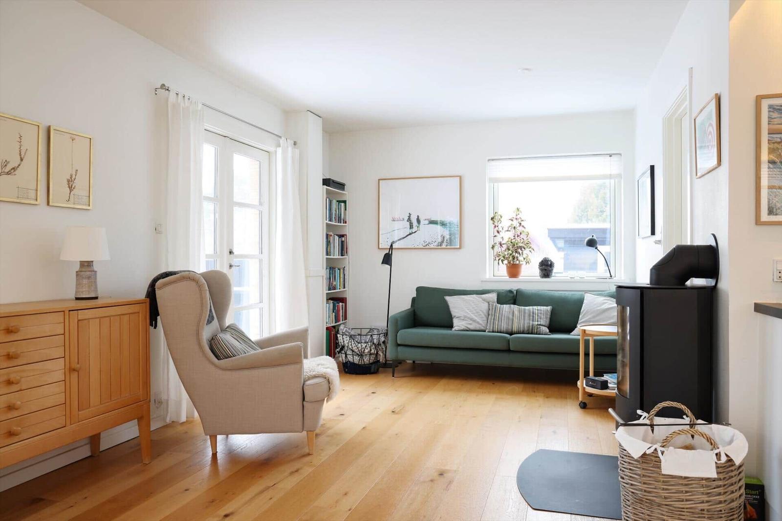Living room with green sofa, wooden floor, and windows. A chair, bookshelf, and wood stove are visible.