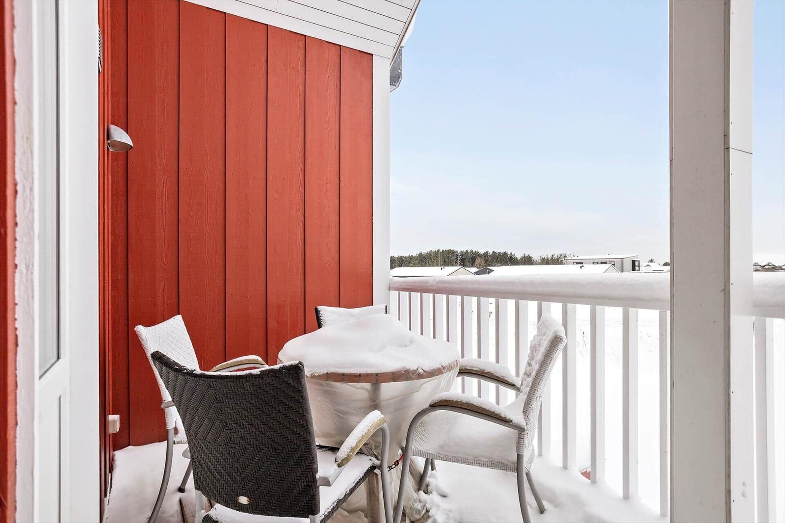 Balcony with table and chairs covered in snow, red wall and white railing.