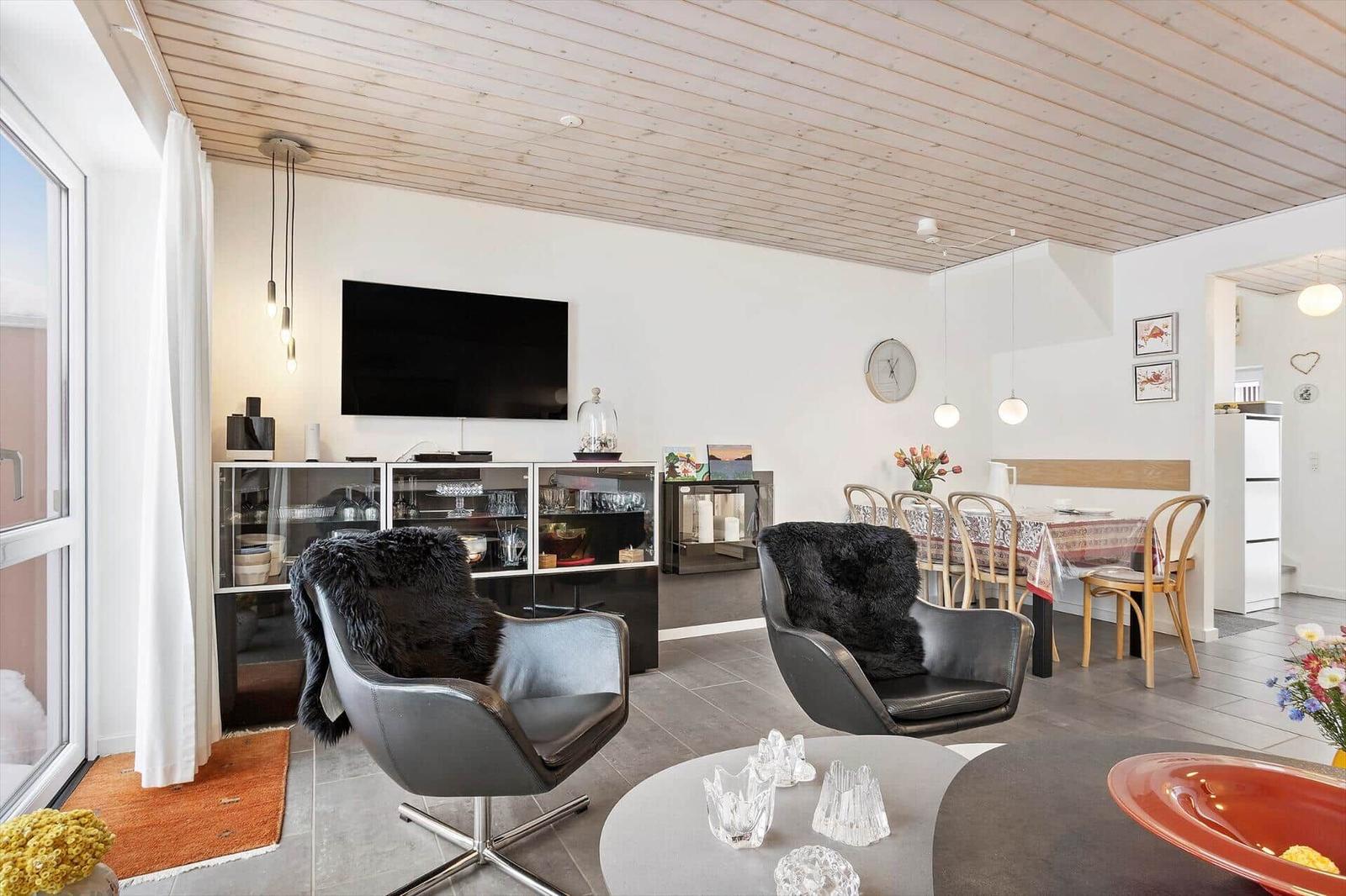 Living and dining area with TV, two chairs, and dining table. Wooden ceiling and modern lighting.