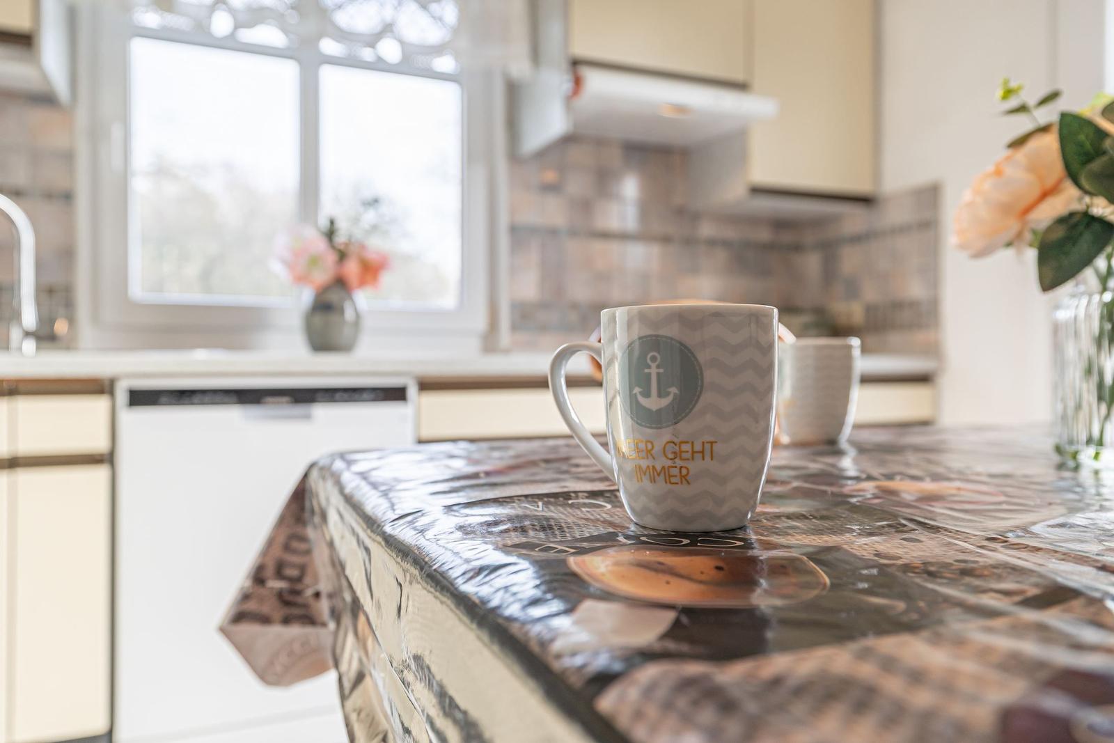 Kücheninsel mit Tischdecke, Tasse und Blumen. Hintergrund: Fenster und Spüle.