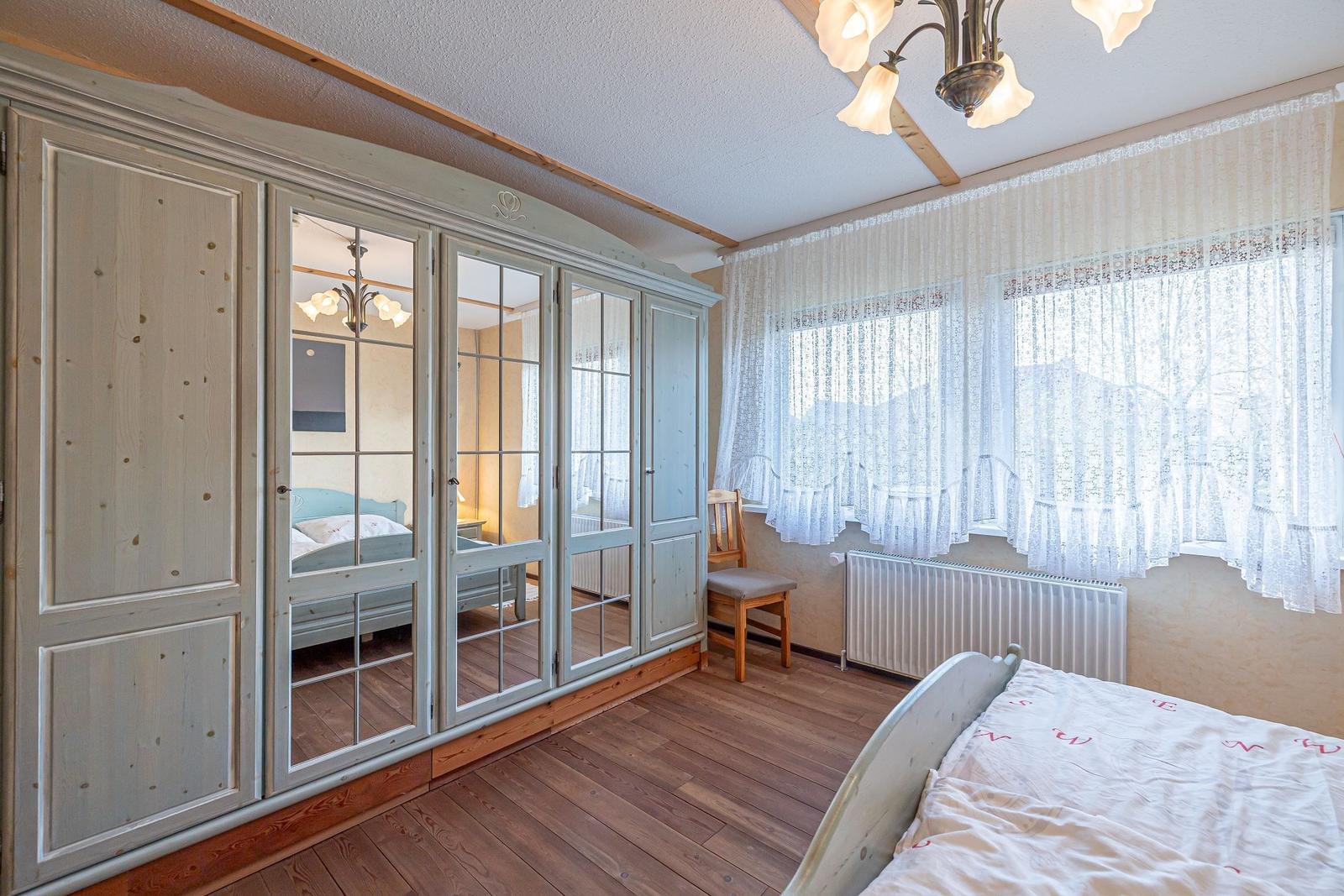Schlafzimmer mit Holzboden, Kleiderschrank mit Spiegeltüren und Fenster mit weißen Vorhängen.