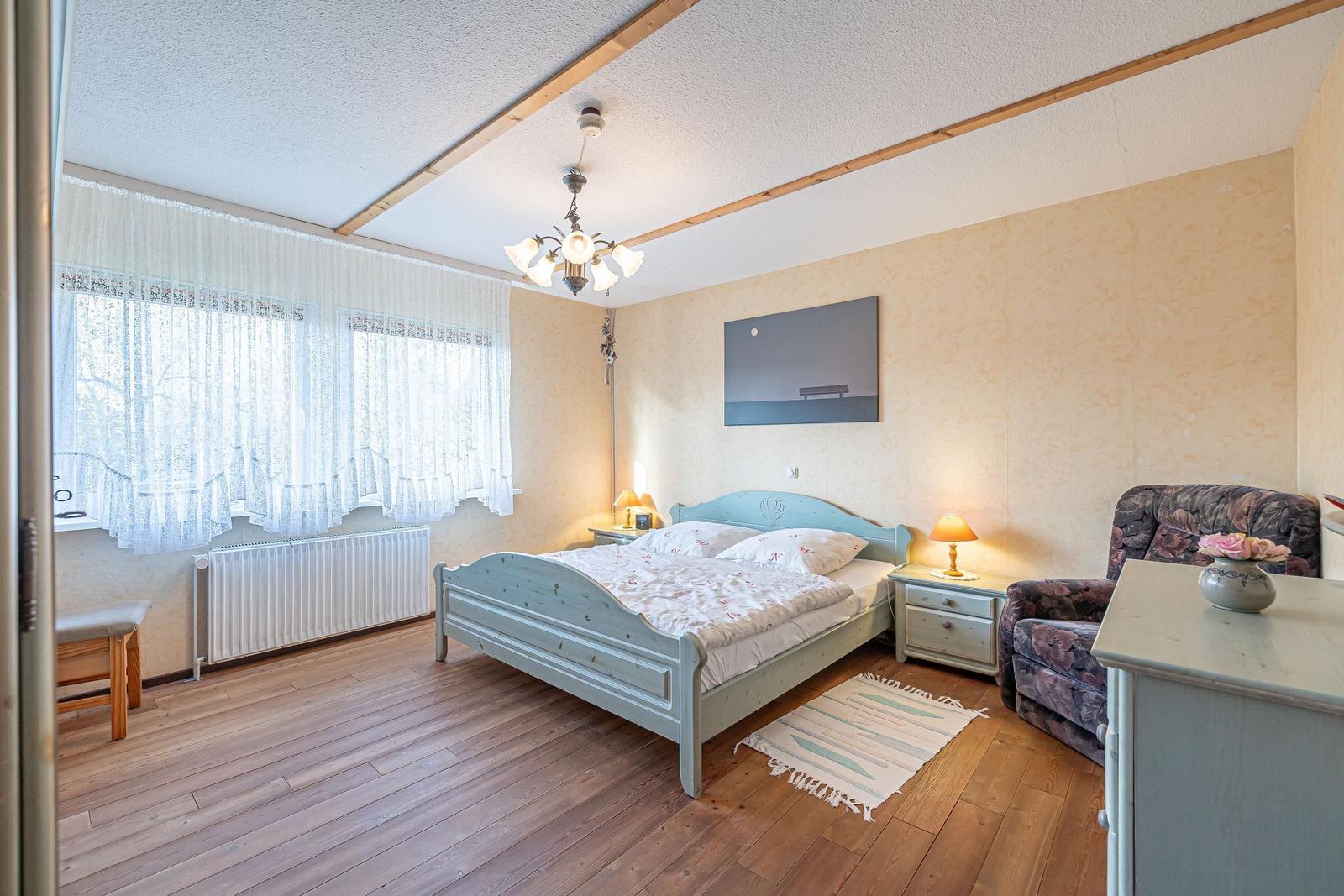 Schlafzimmer mit Bett, Nachttischen, Sessel und Fenster mit Vorhängen.