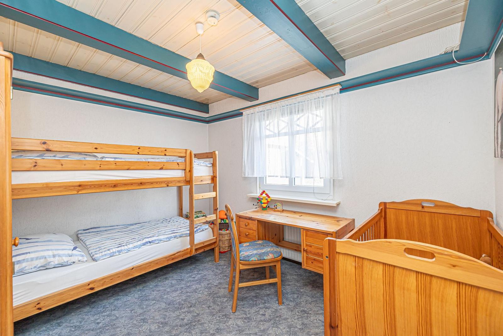 Zimmer mit Doppelstockbett, Schreibtisch und Kinderbett. Holzdecken und Fenster mit Vorhängen.