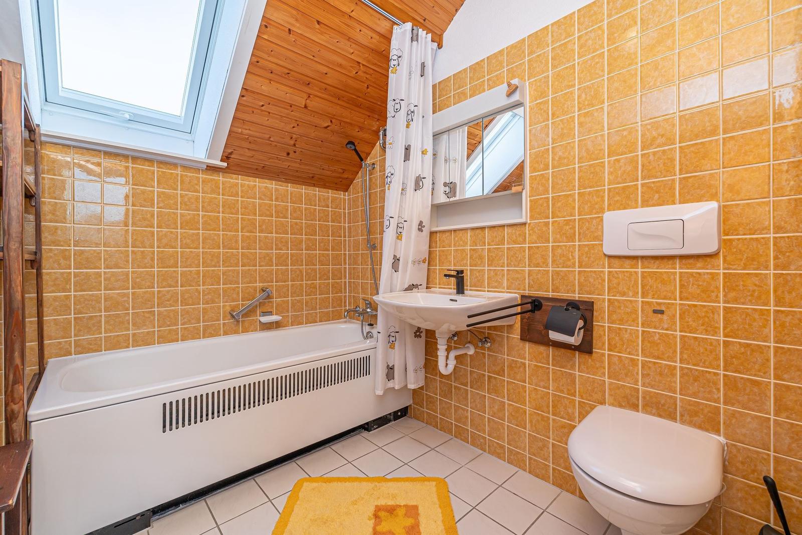 Badezimmer mit Badewanne, Duschvorhang, Waschbecken und WC. Holzdecke und gelbe Fliesen.