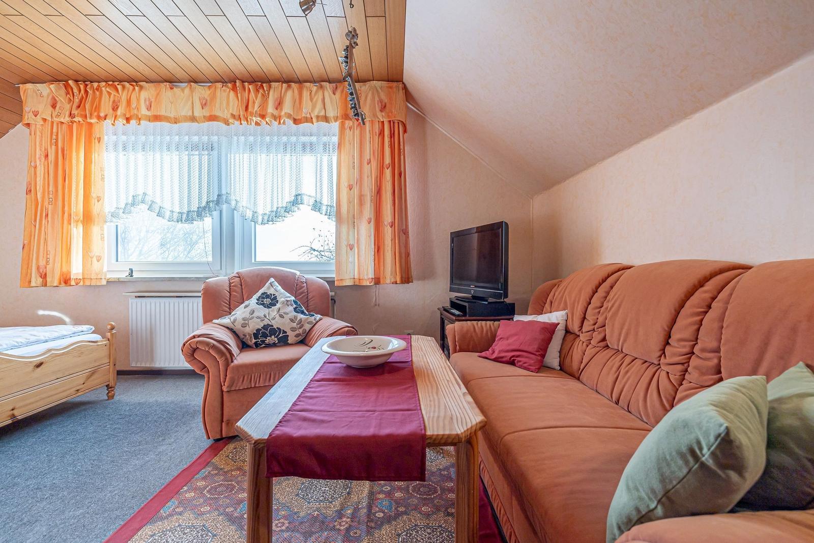 Wohnzimmer mit Sofa, Sessel, Tisch und Fernseher. Fenster mit Vorhängen und Bett im Hintergrund.