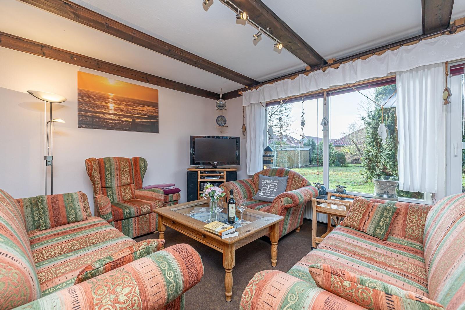 Wohnzimmer mit Sofa, Sessel und TV. Durchblick auf Garten durch große Fenster.