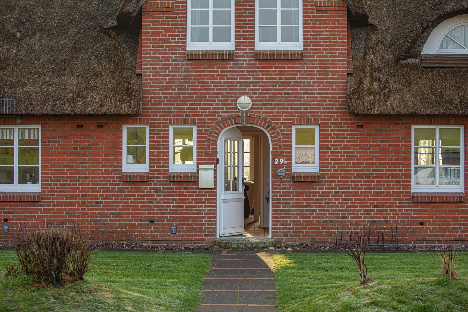 Rotes Backsteinhaus mit Strohdach und weißer Eingangstür. Hausnummer 29e. Gras und Steinstiege.