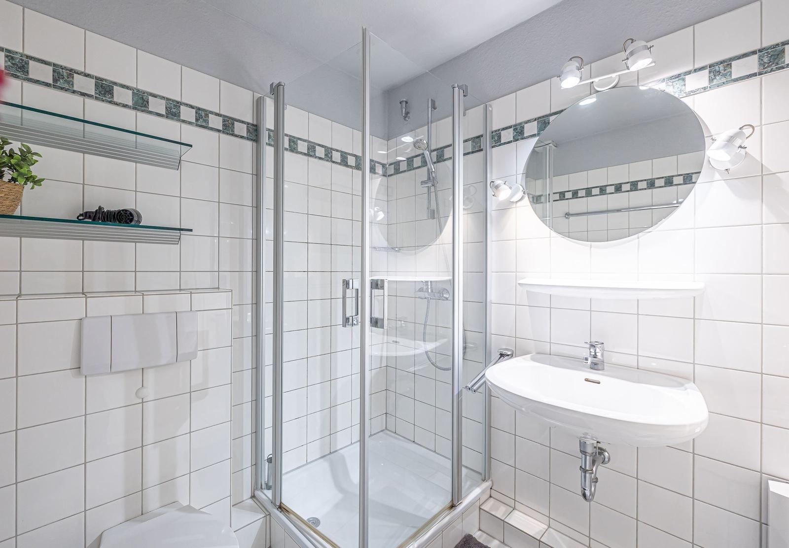 Badezimmer mit Dusche, Waschbecken und Spiegel. Weiße Fliesen und Glaswand.