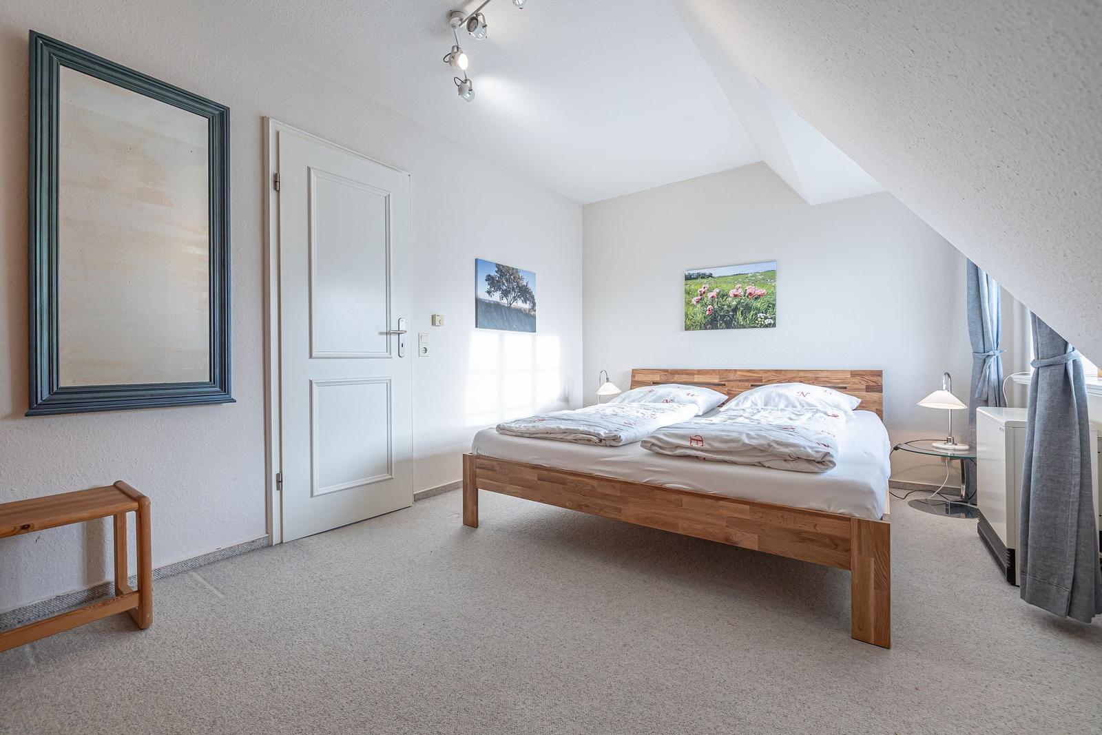 Schlafzimmer mit Dachschrägen, Holzbett, Spiegel und Fenster mit Vorhängen.