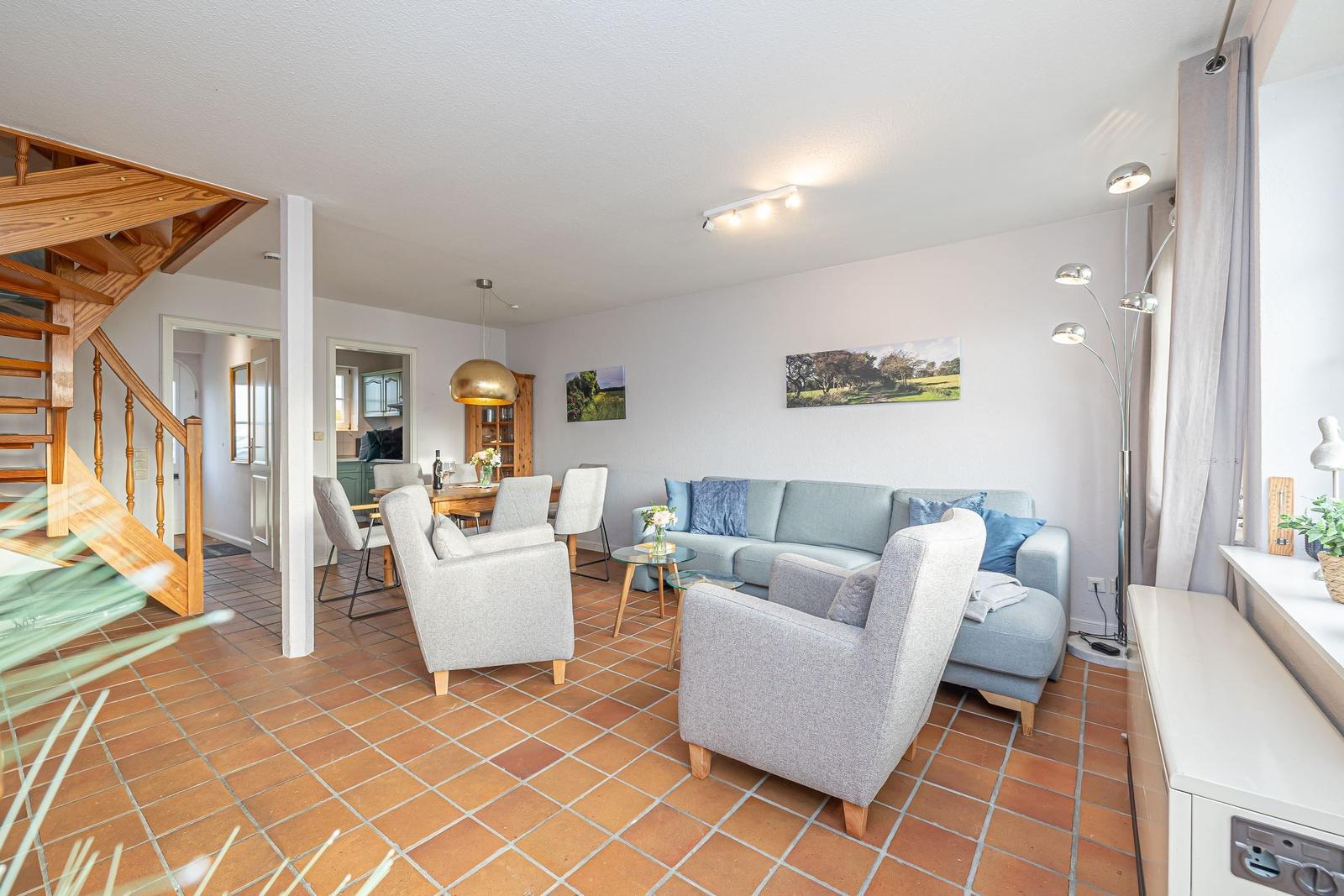 Wohn- und Essbereich mit Sofa, Sesseln und Holztreppe. Terrakottaboden und Wandbilder.