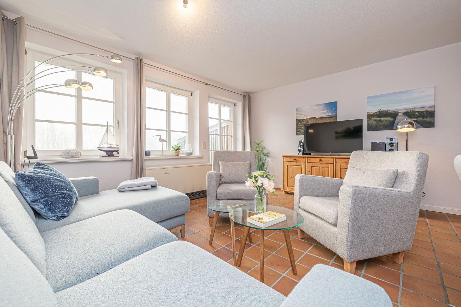 Wohnzimmer mit Sofa, zwei Sesseln, Glas-Tisch und Fernseher. Terrakotta-Boden und Fenster mit Vorhängen.