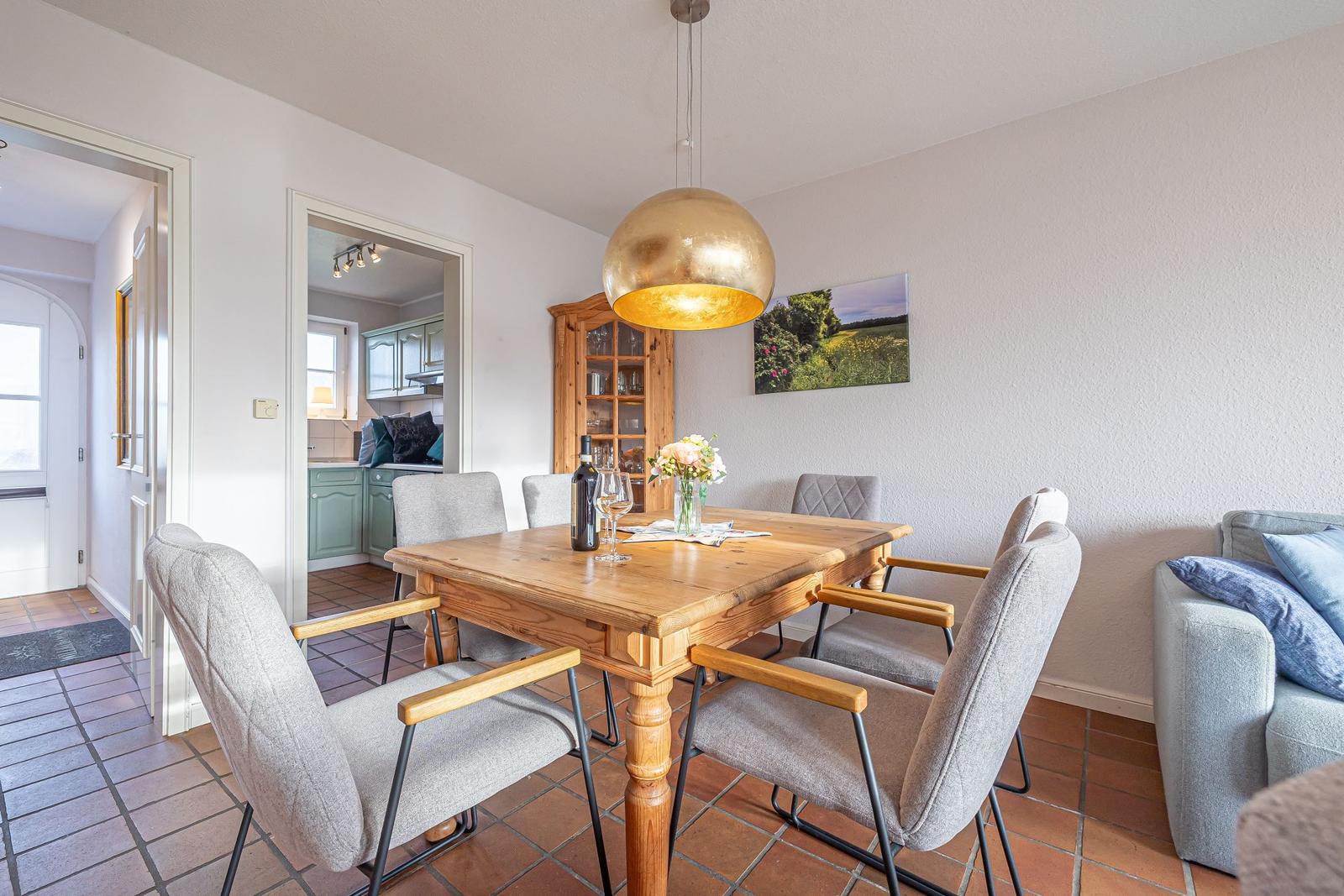 Essbereich mit Holztisch, Stühlen und goldener Deckenleuchte. Küche und Wohnbereich sind sichtbar.