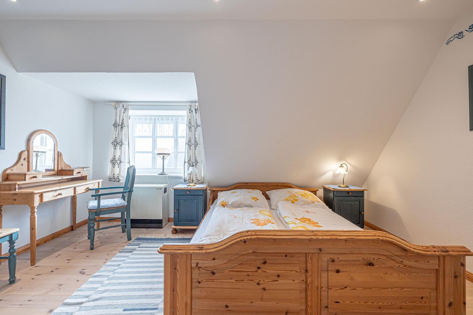 Schlafzimmer mit Holzbett, Schreibtisch und Fenster. Holzfußboden und blaue Nachttische.