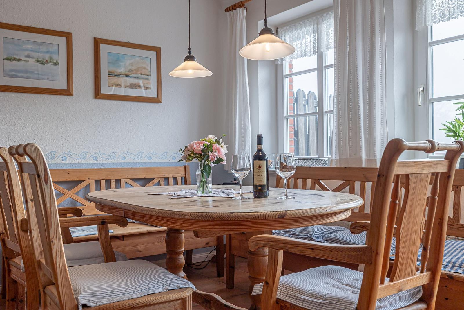 Esszimmer mit Holztisch, Stühlen und Fenster. Auf dem Tisch steht eine Flasche Wein und Blumen.