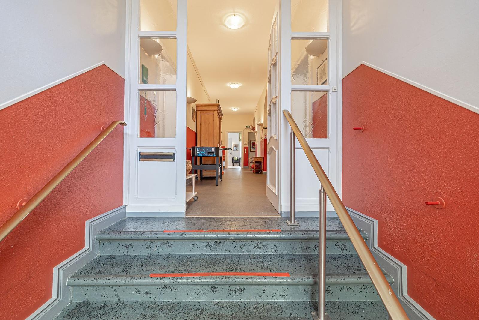 Treppenhaus mit roten Wänden und grauen Stufen führt in einen Flur mit Möbeln.