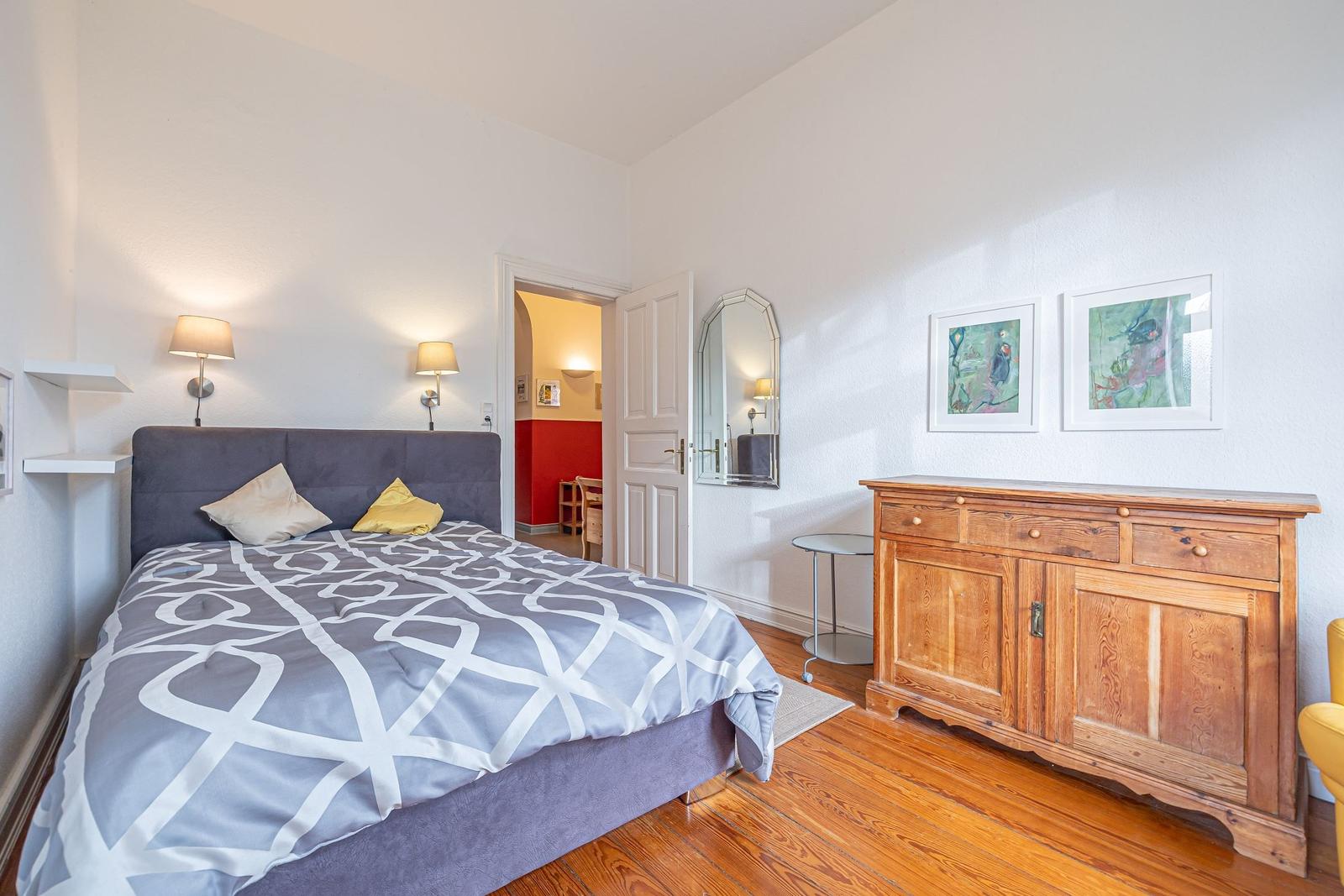 Schlafzimmer mit Bett, Holzschrank und Spiegel. Tür führt in angrenzenden Raum.