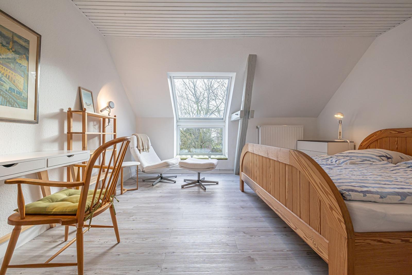 Schlafzimmer mit Holzbett, Schreibtisch und Sessel unter Dachschrägen.