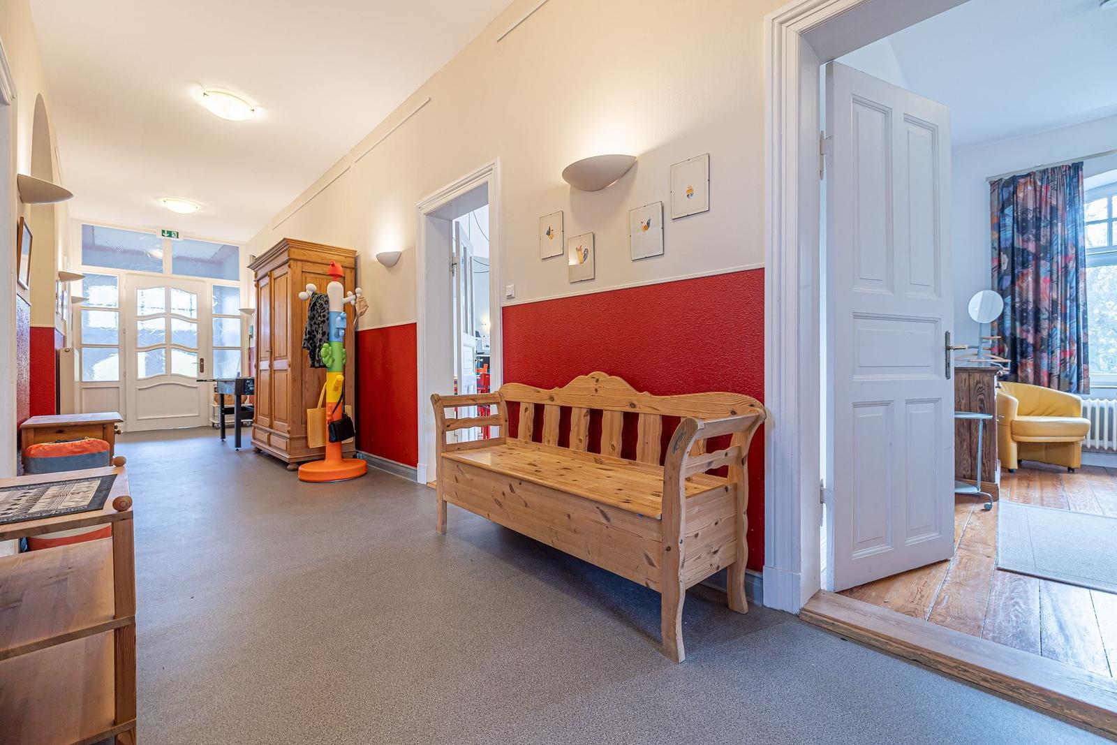 Hallenflur mit Holzbank, roter Wand und Türöffnung zu einem Zimmer.