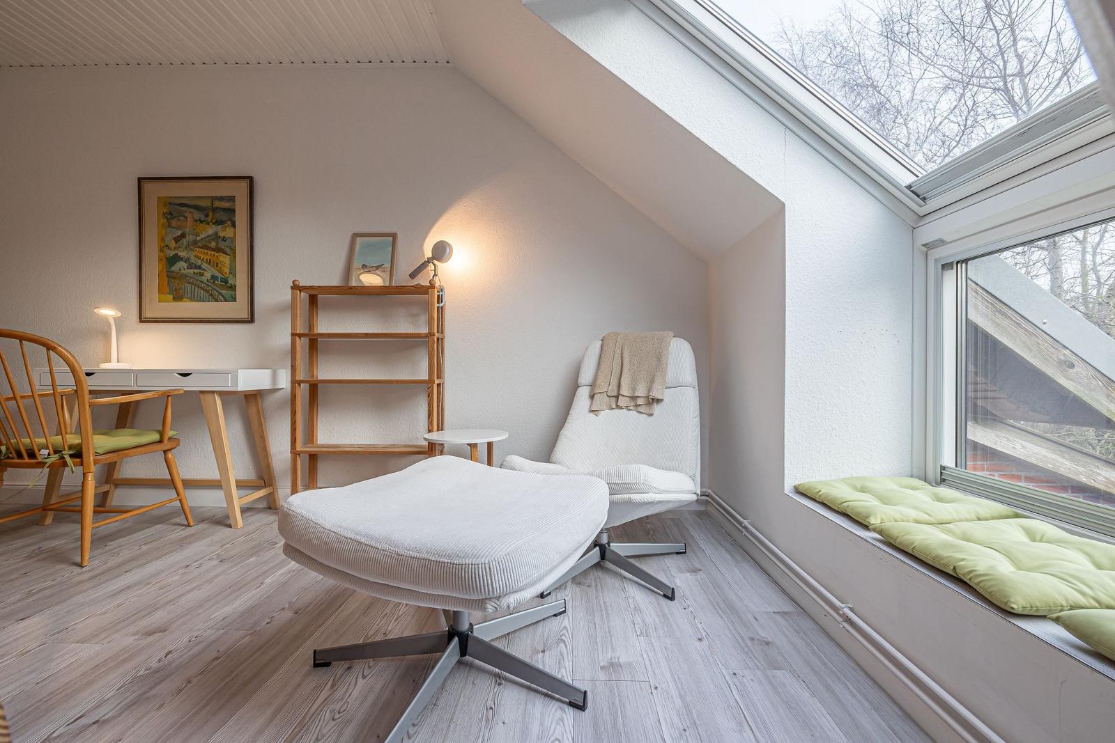 Ein Schlafzimmer mit Dachfenster, Schreibtisch, Stuhl und weißem Sessel mit Fußstütze.