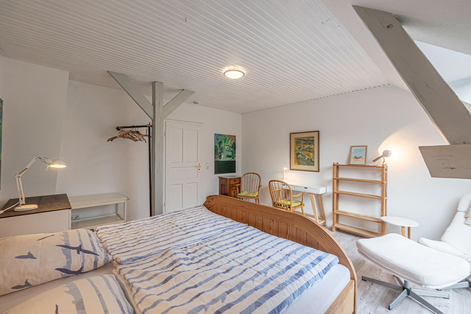 Schlafzimmer mit Bett, Schreibtisch, Stühlen und Regal. Holzbalken an der Decke.