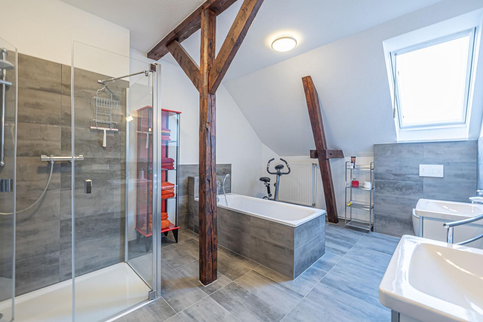 Badezimmer mit Dusche, Badewanne, Toilette und Fahrradtrainer unter Holzträgern.
