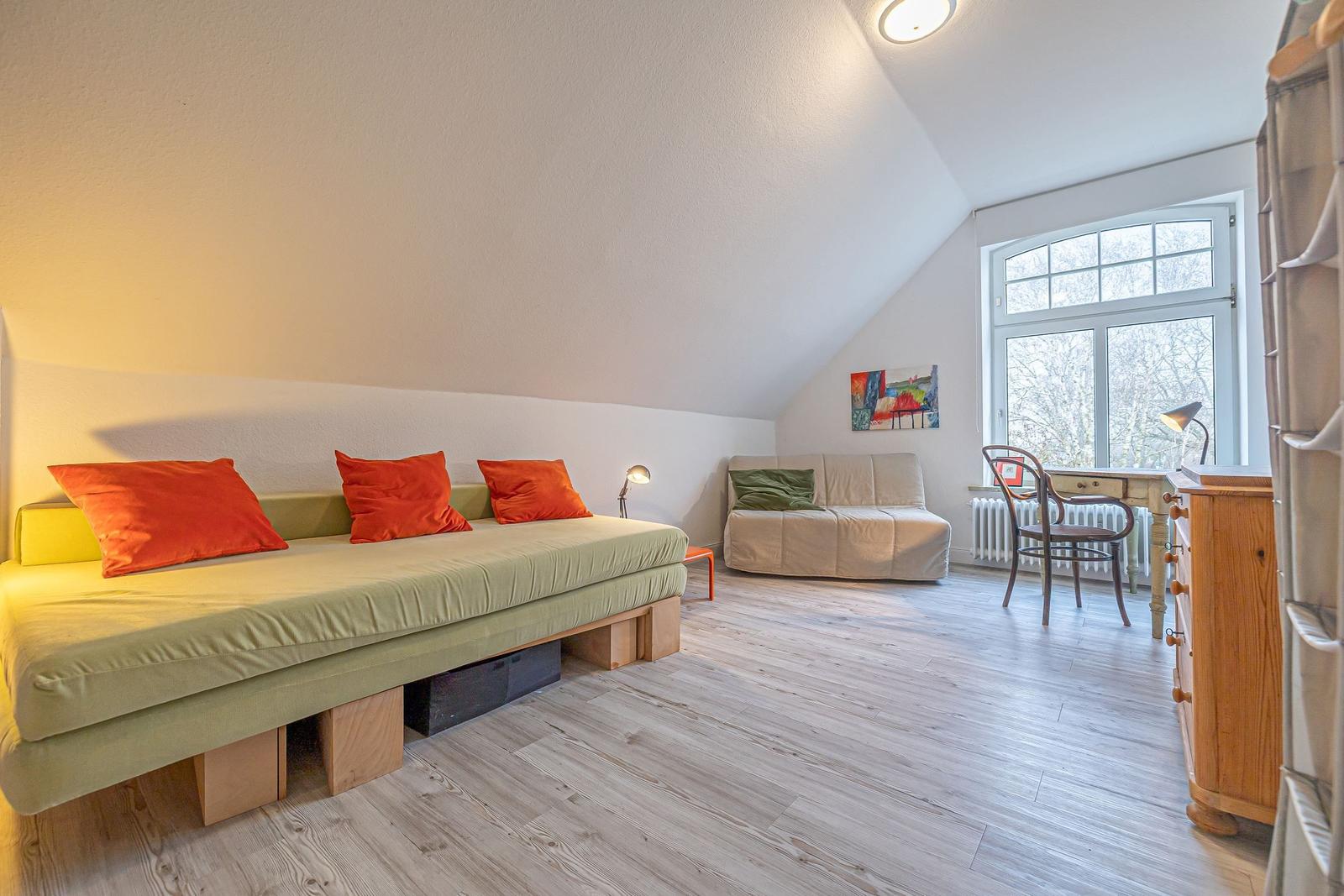 Ein Zimmer mit Schlafcouch, kleiner Sitzbank, Schreibtisch und großem Fenster.