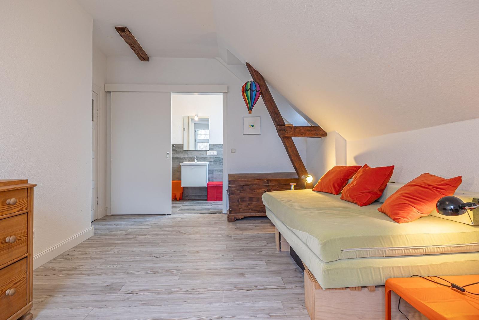 Schlafzimmer mit Dachschrägen, Bett mit orangefarbenen Kissen und Blick in die Badewanne.